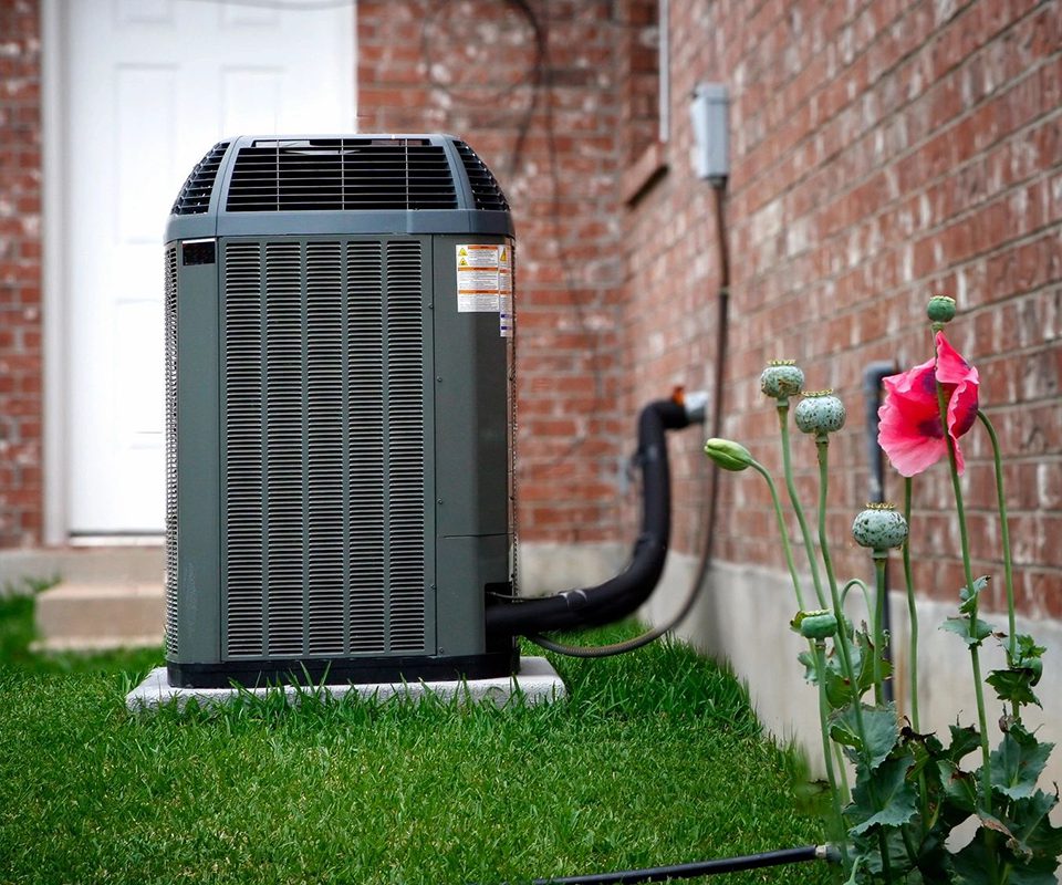 An outdoor air conditioning unit sits next to a brick house with a white door in the background and pink flowers growing in the grass nearby.