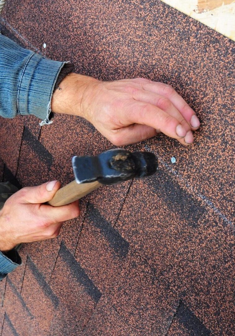 Person in a denim shirt nailing a shingle onto a roof with a hammer.